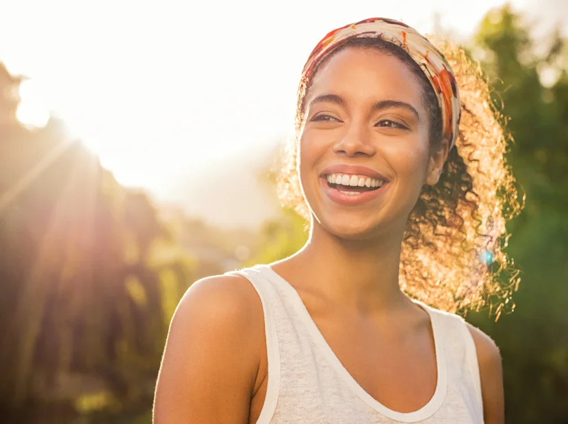 young adult female with curly hair in a headband smiling against a sunset in background